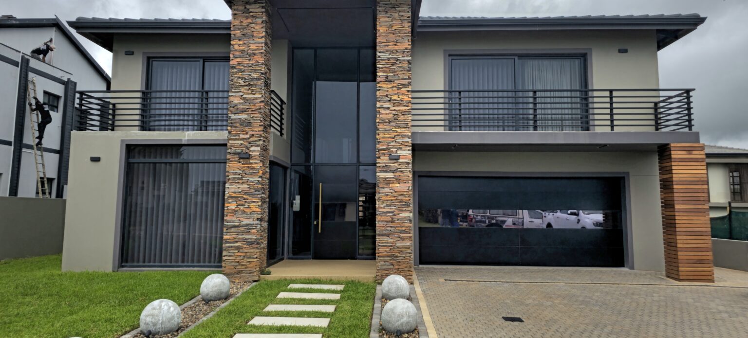 Double-storey home with black aluminium garage door and glass entrance by Quality Design Doors